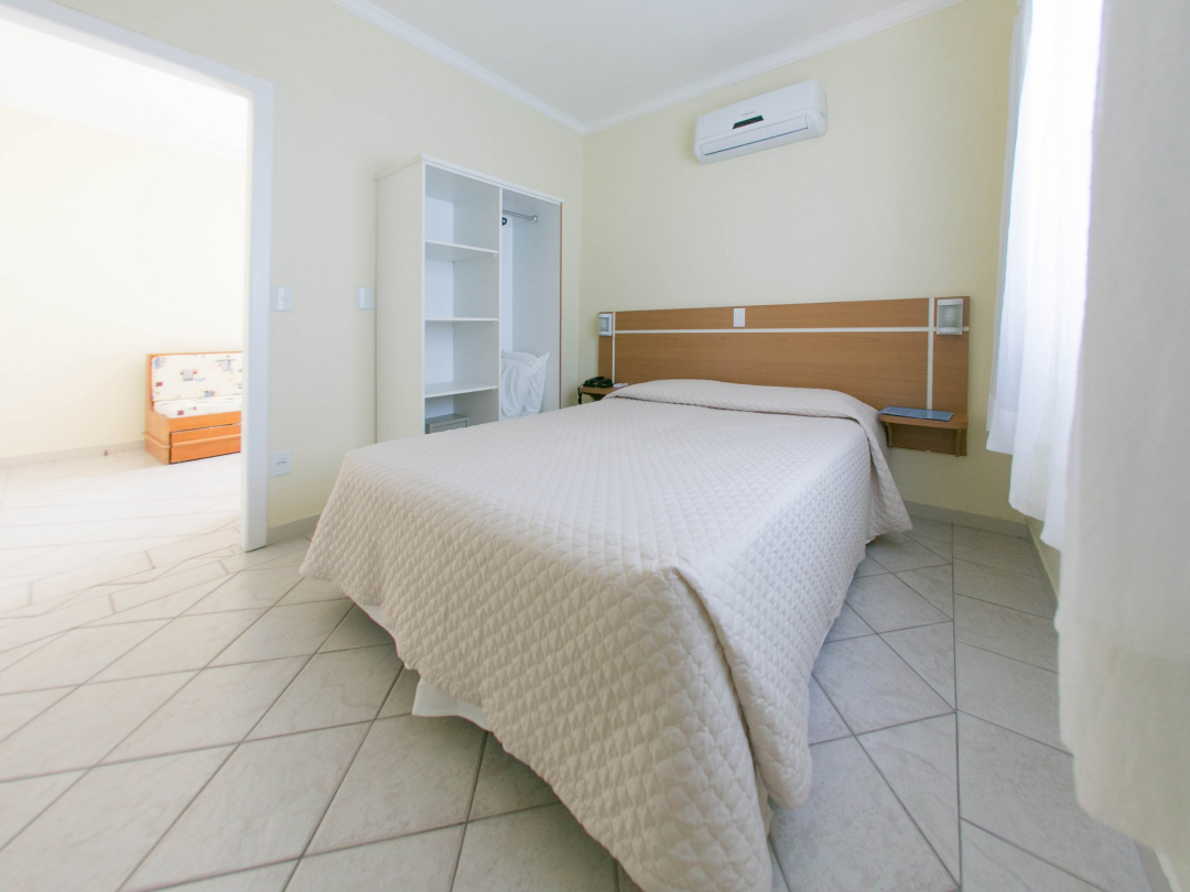 Foto ampla do quarto do apartamento família com cozinha e varanda no Bombinhas Praia Apart Hotel II mostrando a cama e o ar condicionado.
